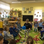 Dzieci siedzą w kole na krzesełkach, dziewczynka w środku trzyma niebieski balon